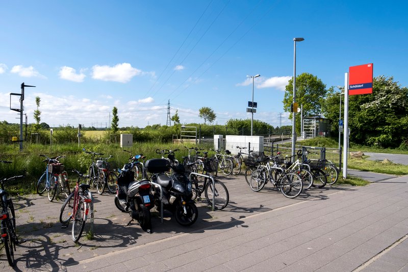 Fahrräder und ein Roller stehen an einem sonnigen Bahnhof mit dem Schild 'Schülldorf'.