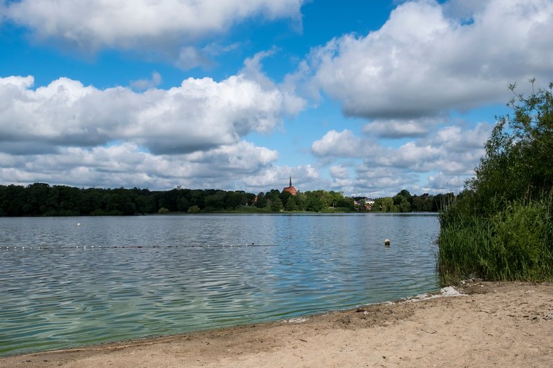 Ein Sandstrand am Bordesholmer See mit Blick auf das Wasser und eine Kirche im Hintergrund. Der Himmel ist bewölkt mit einigen blauen Stellen.