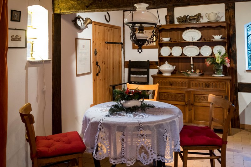 Gemütliche Stube mit Holzmöbeln, Tisch mit Spitzendecke und Dekoration, Geschirrschrank im Hintergrund.