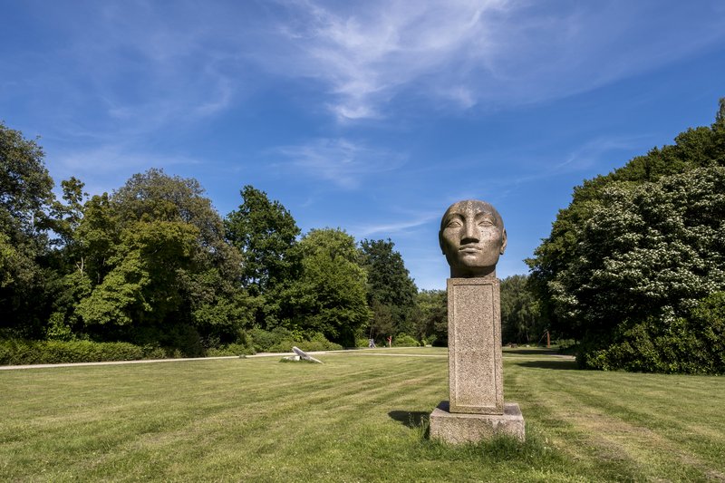Große Steinskulptur eines Kopfes auf einem Sockel im Nortorfer Park, umgeben von grünen Bäumen und Rasen unter blauem Himmel.