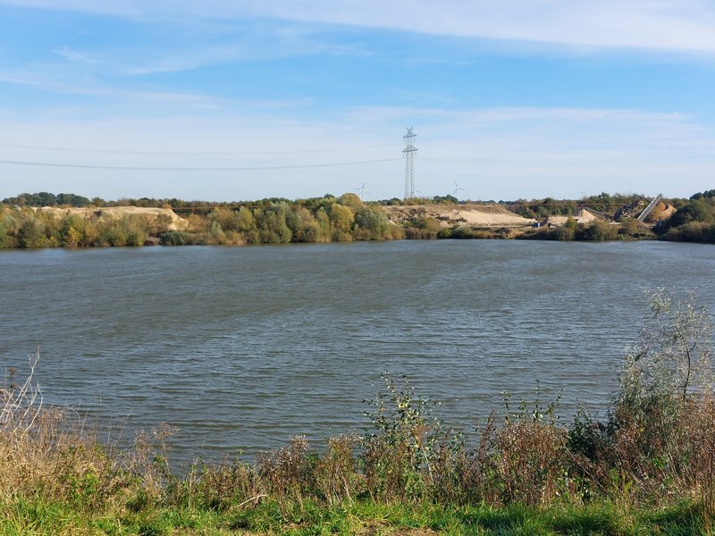 Ein See mit bewachsenen Ufern und einem Hügel im Hintergrund, darüber ein blauer Himmel mit einigen Wolken.
