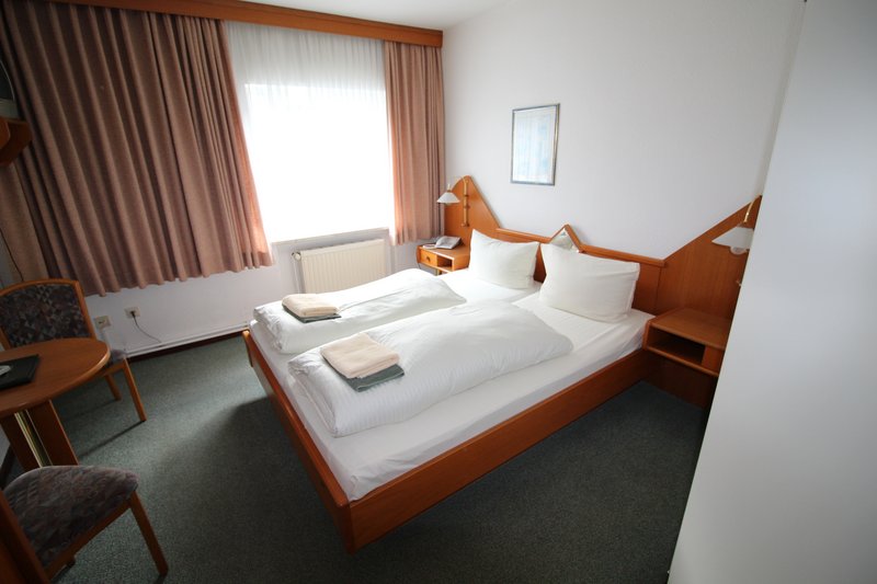 Ein Hotelzimmer mit einem Doppelbett, weißen Bettlaken und Handtüchern. Ein Fenster mit Vorhängen und ein kleiner Tisch mit Stuhl sind ebenfalls zu sehen.