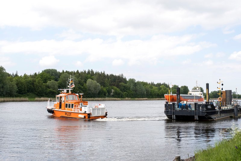 Ein orangefarbenes Lotsenboot fährt auf einem Fluss, während ein größeres Schiff im Hintergrund zu sehen ist. Am Ufer sind Bäume und Gras zu erkennen.