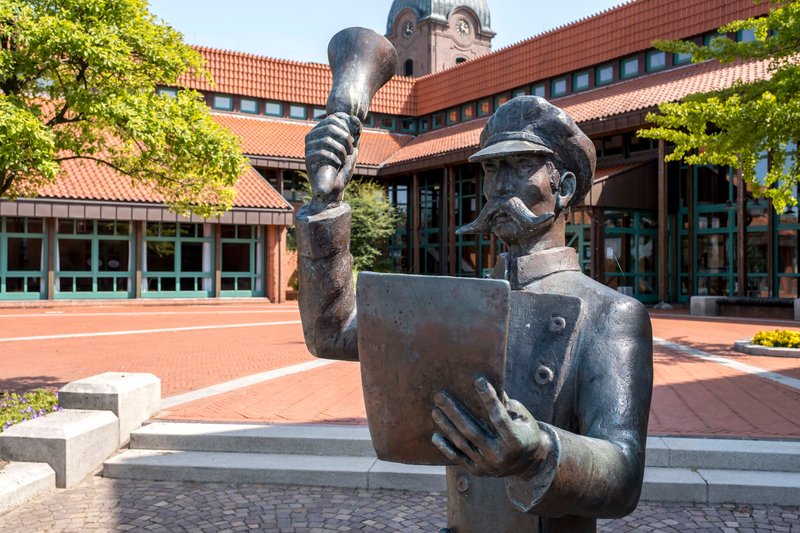 Bronzestatue eines Mannes mit Megafon und Papier vor einem Gebäude mit rotem Ziegeldach.