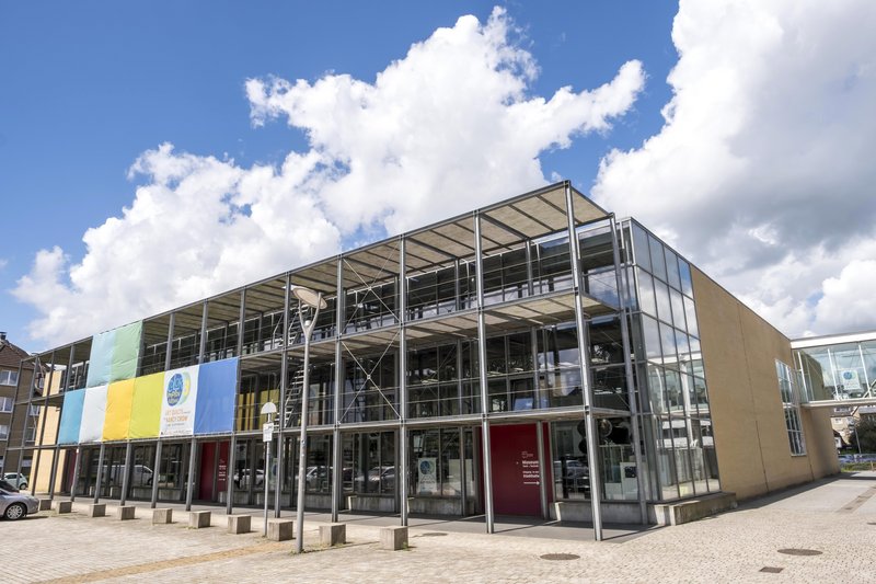Modernes Gebäude des Museums Tuch & Technik in Neumünster mit Glasfassade und bunten Bannern.
