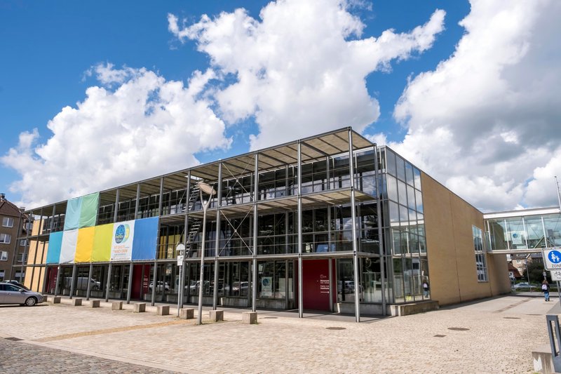 Außenansicht des Museums Tuch & Technik in Neumünster mit moderner Glasfassade und bunten Bannern.