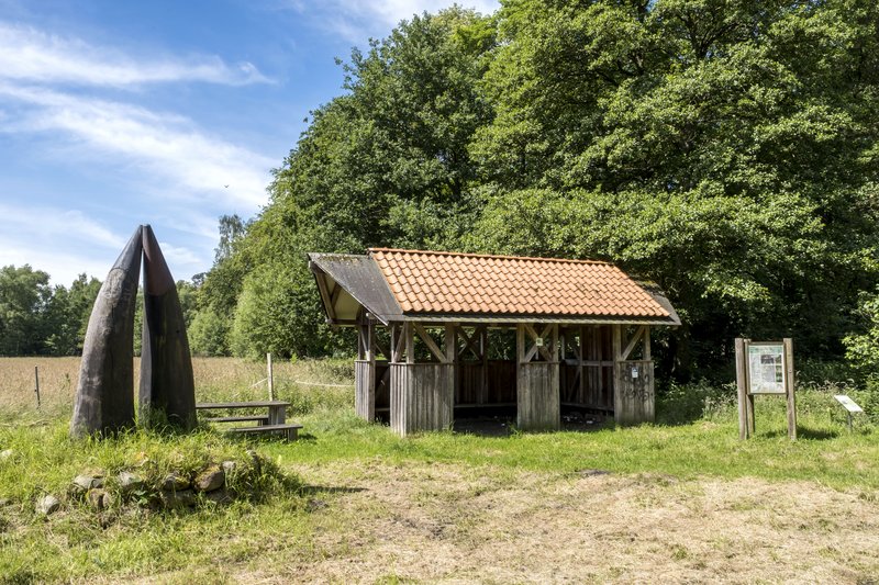 Ein Rastplatz mit einem hölzernen Unterstand und einer Skulptur aus zwei großen Hörnern auf einer Wiese, umgeben von Bäumen.